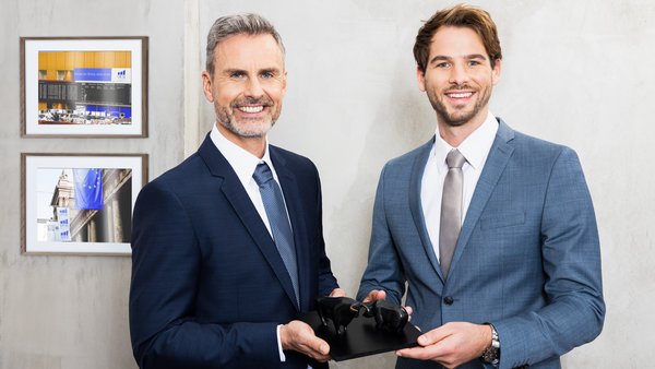 Két öltönyös férfi együtt tart egy bikát és medvét ábrázoló szobrot – ez szimbolikusan a pénzügyi piacokat és a partnerségi együttműködést jelképezi.