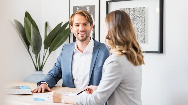 A kék zakós férfi barátságosan beszélget egy világos blézeres nővel az íróasztalnál.