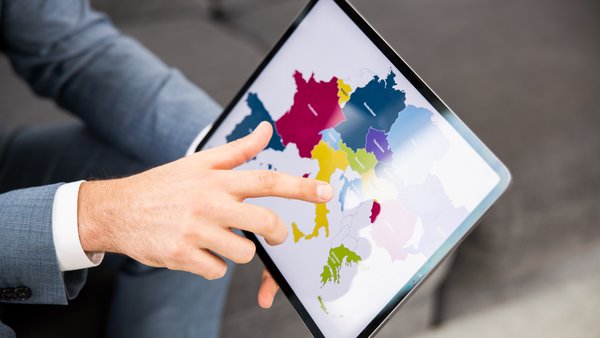 Egy személy kezében egy táblagépet tart, amelyen Európa térképe látható, és ujjal mutat a térképre.